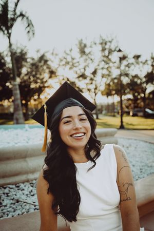 Graduate in cap and gown smiles brightly during outdoor sunset photoshoot.