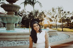 Graduate in white dress and academic cap poses by fountain during golden hour.