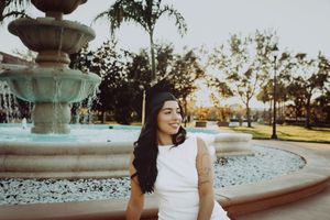 Graduate in white dress poses near outdoor fountain at sunset.