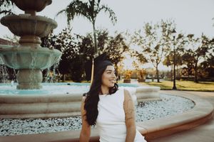 Graduate in white sleeveless dress poses by fountain during golden hour.