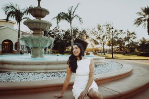 Graduate in white dress sitting by ornate fountain with palm trees in background.
