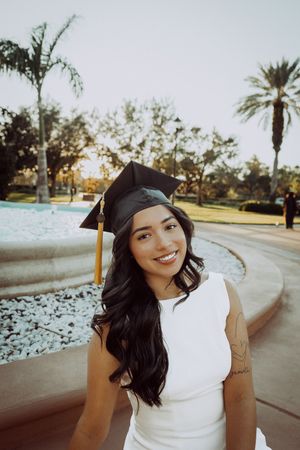 Graduate in cap and gown smiling by fountain with palm trees in background.