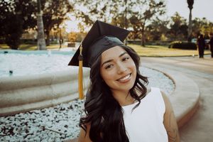 Graduate in academic regalia smiles during outdoor portrait session near palm trees.
