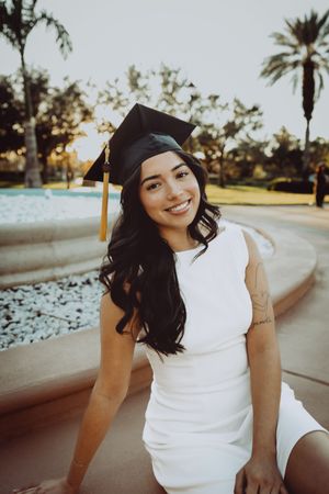Graduate in cap and gown sits by decorative fountain in palm tree setting.