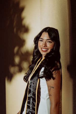 Graduate wearing black graduation stole and cap poses against neutral backdrop.