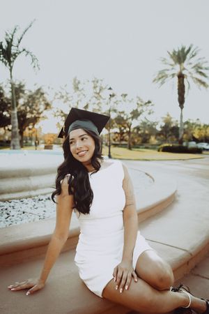 Graduate in white dress and cap relaxing by circular fountain.