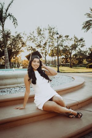 Graduate in cap and white dress posing on fountain steps at sunset.