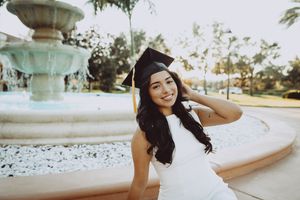 Graduate relaxes by ornate stone fountain during golden hour photoshoot.