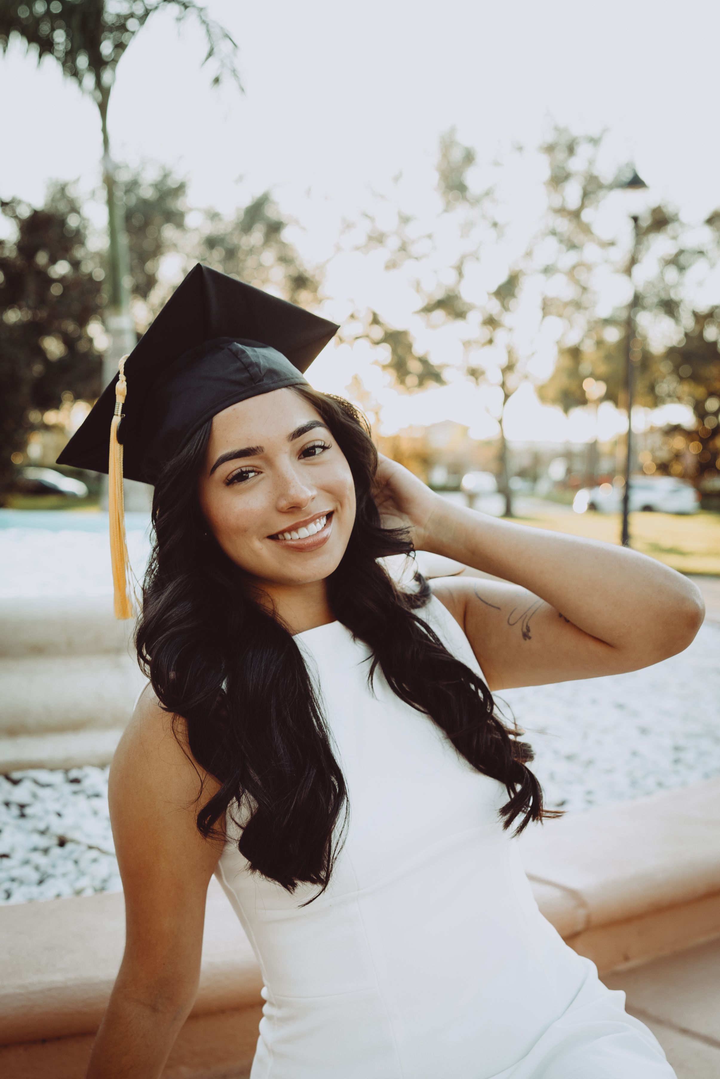 Graduate in white dress and mortarboard cap poses outdoors during sunset photoshoot.