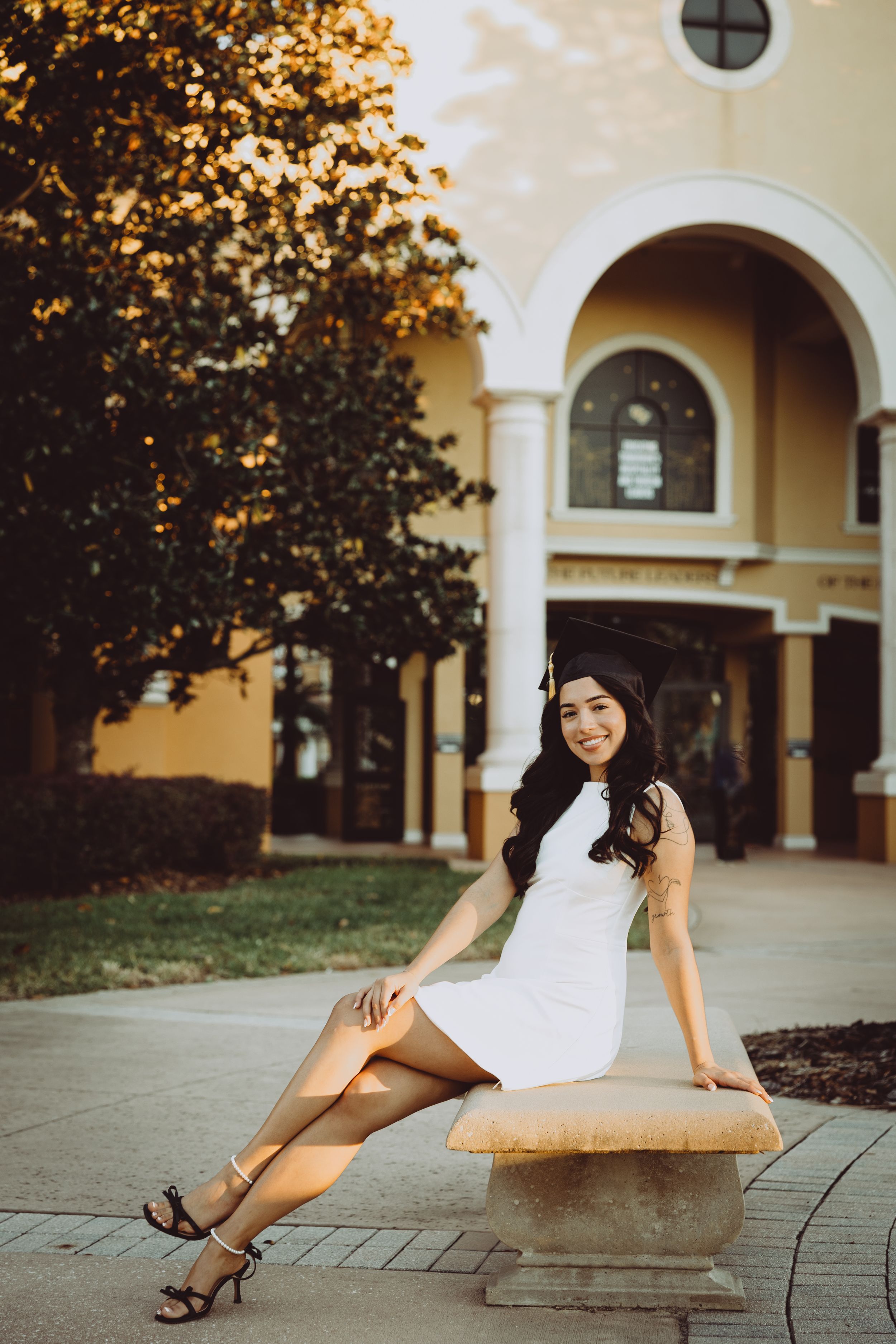 A person in a white dress poses gracefully on a stone bench against a Spanish colonial style building with arches.