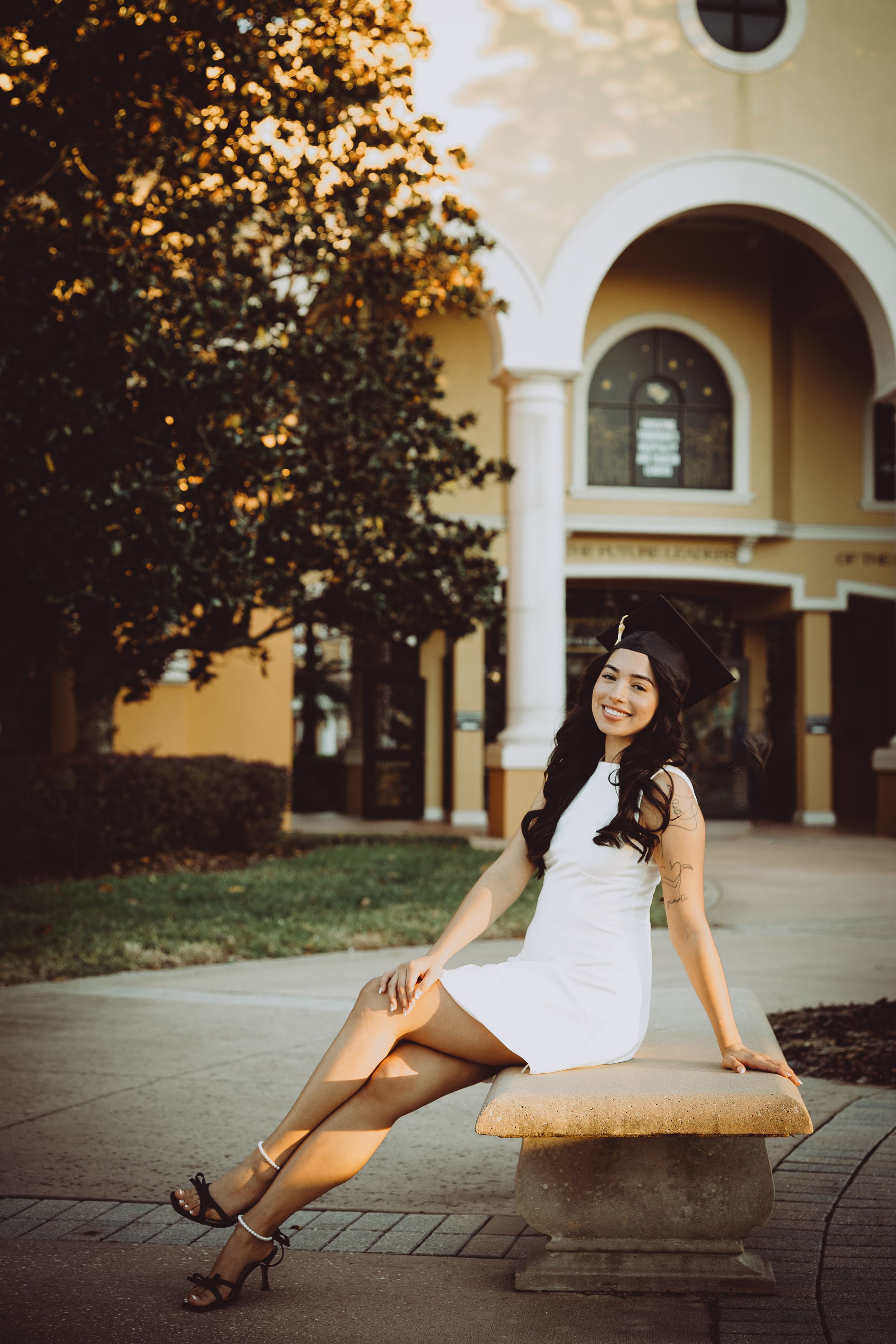 Graduate posing in a white dress and heels against architectural columns at sunset.