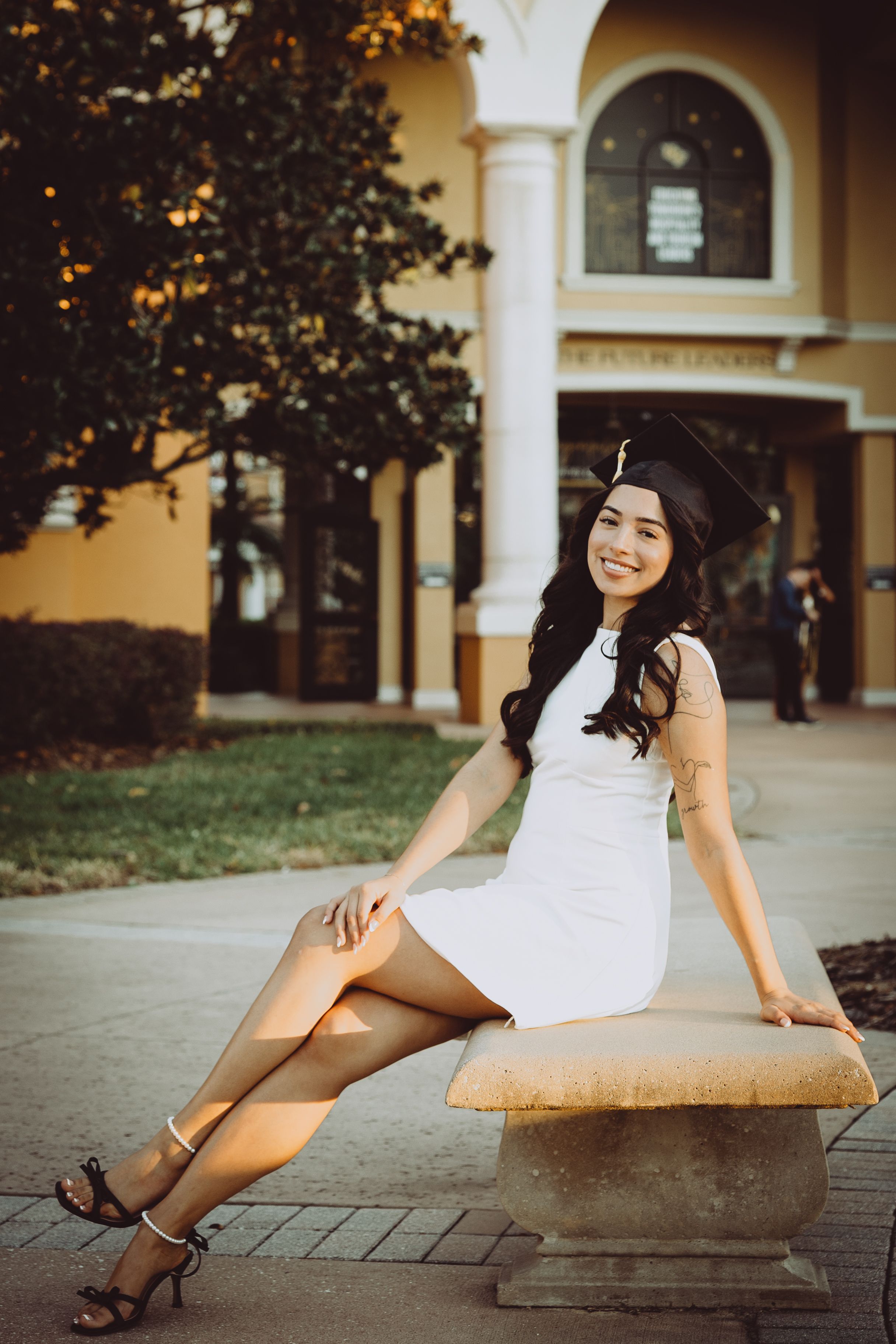 Graduate in white dress poses playfully on outdoor campus grounds with arched architecture.
