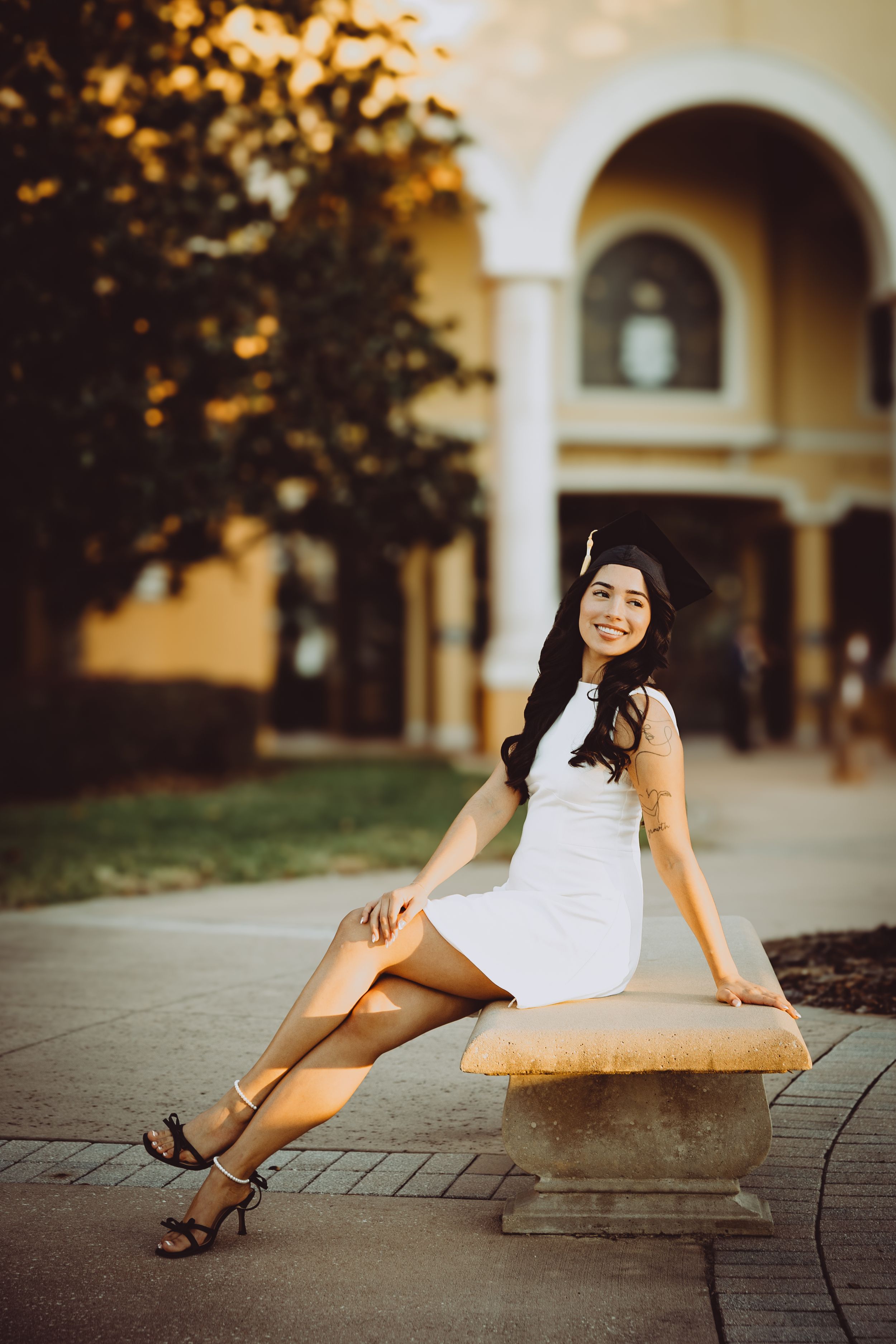 A person in a white dress poses gracefully on a brick walkway near arched columns during golden hour.