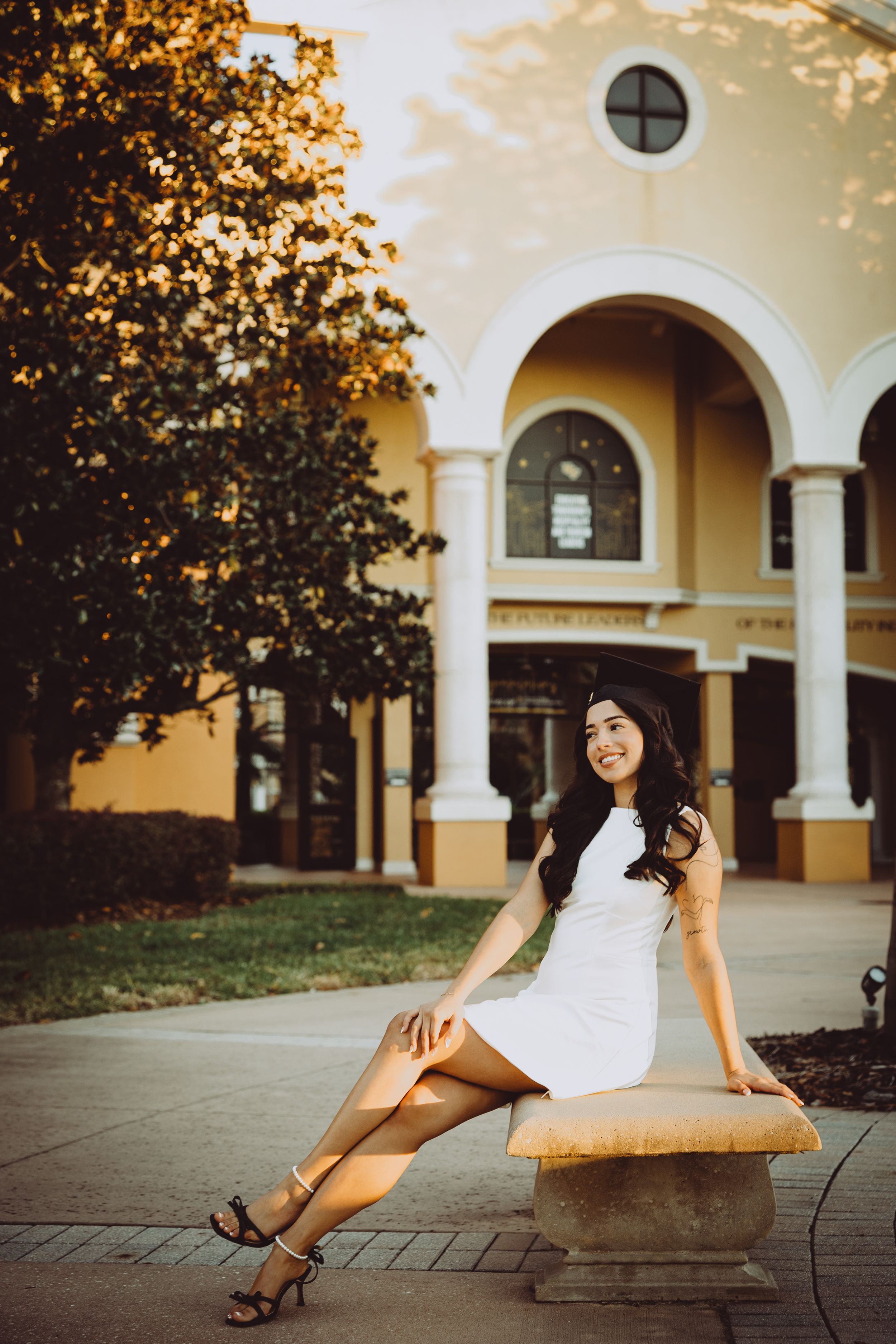 Graduate in a white dress poses elegantly on a stone bench against Spanish colonial architecture at sunset.