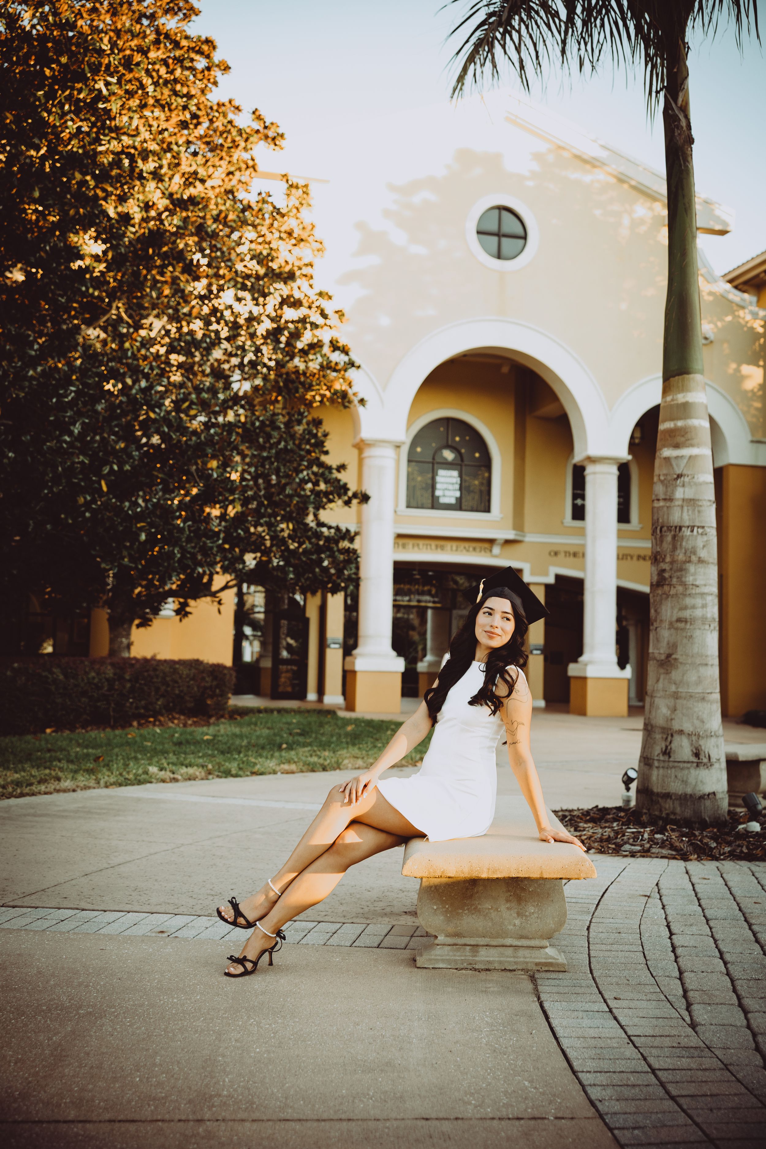 A person in a white dress poses on a bench outside a Mediterranean-style building with arches and palm trees.