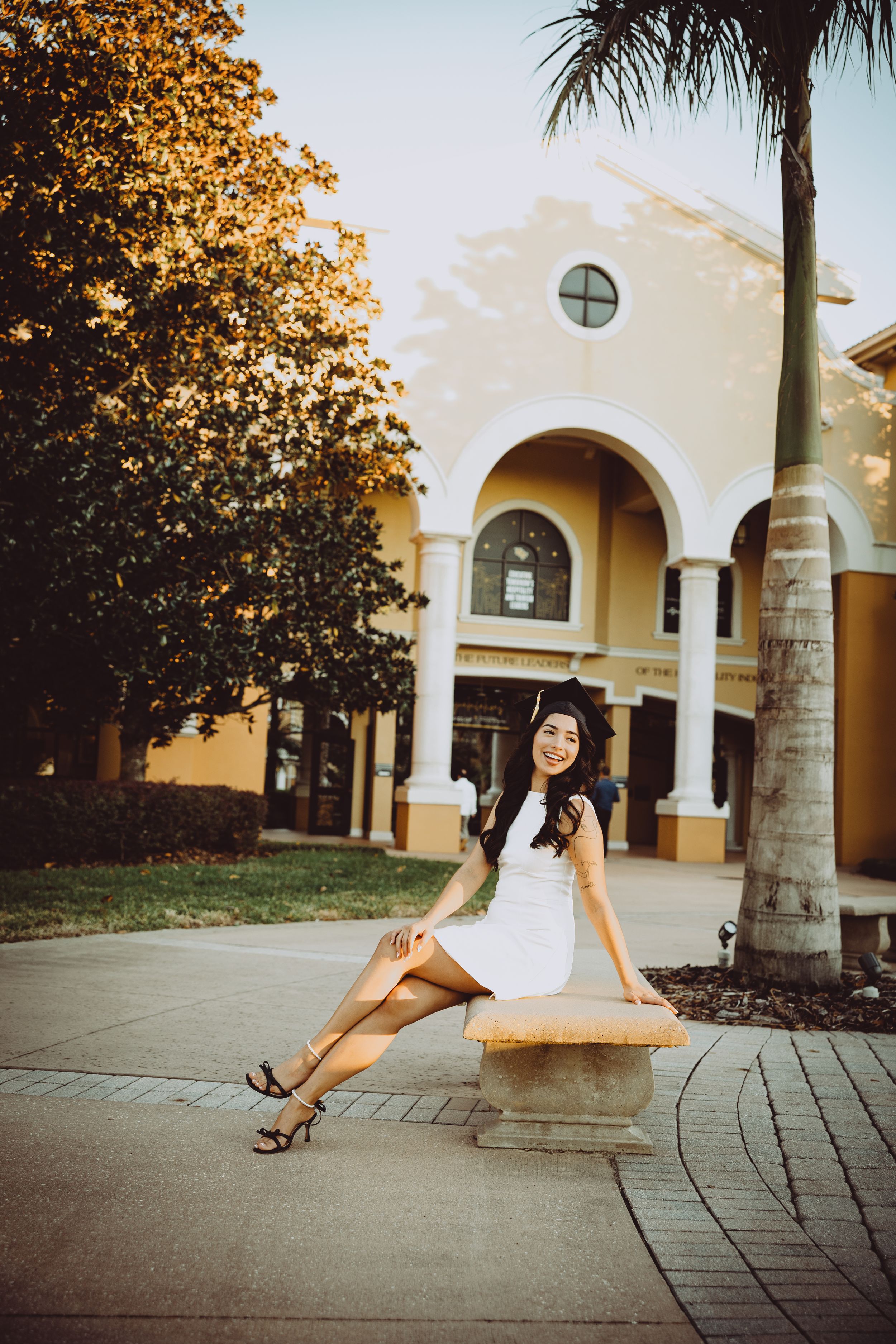 Graduate in white dress poses near palm tree and Spanish colonial architecture.