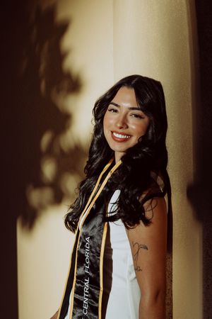 Graduate in black and white graduation stole smiles warmly in natural lighting with shadows cast on wall.