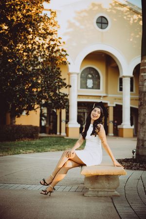 Graduate poses on concrete path near Spanish-style building with arched columns.