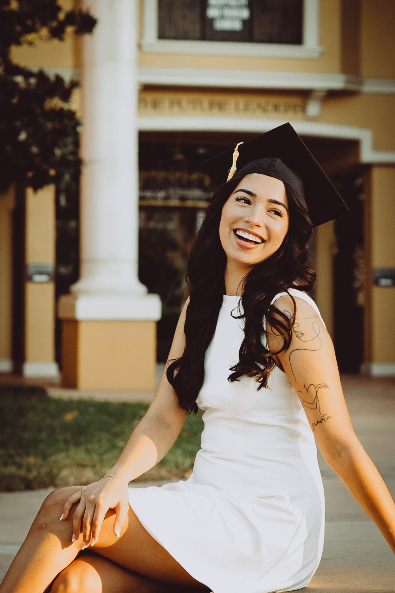 Graduate in white dress sits on ground with graduation cap while smiling during outdoor photo session.