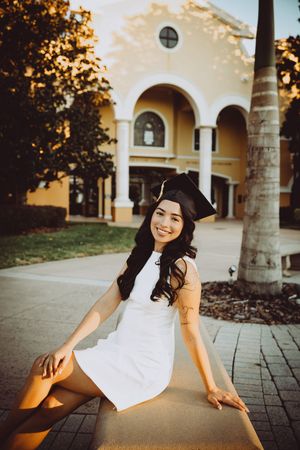 Graduate in white dress sits on concrete path during outdoor photo session.