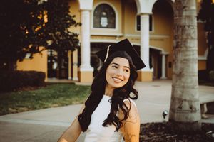 Graduate in black cap and gown stands outdoors near building columns during golden hour photography session.