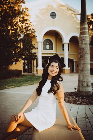 Graduate in cap and white dress sits casually on stone bench with arched walkway in background.