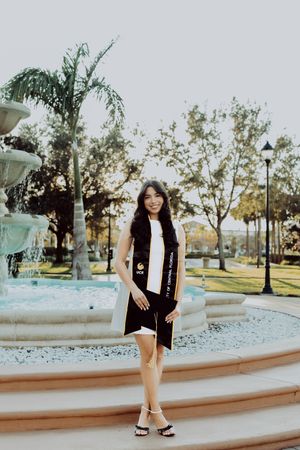 Graduate in cap and gown poses in a park setting with palm trees and curved walkway at sunset.