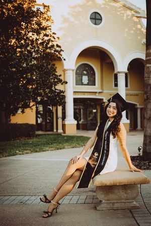 Graduate in cap and gown poses near Mediterranean-style building with arches at sunset.