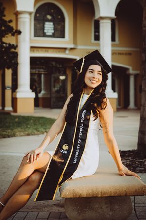 Graduate in white dress poses with graduation sash against yellow building with arches.