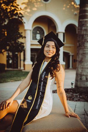 Graduate wearing academic stoles poses on campus grounds near arched walkway.
