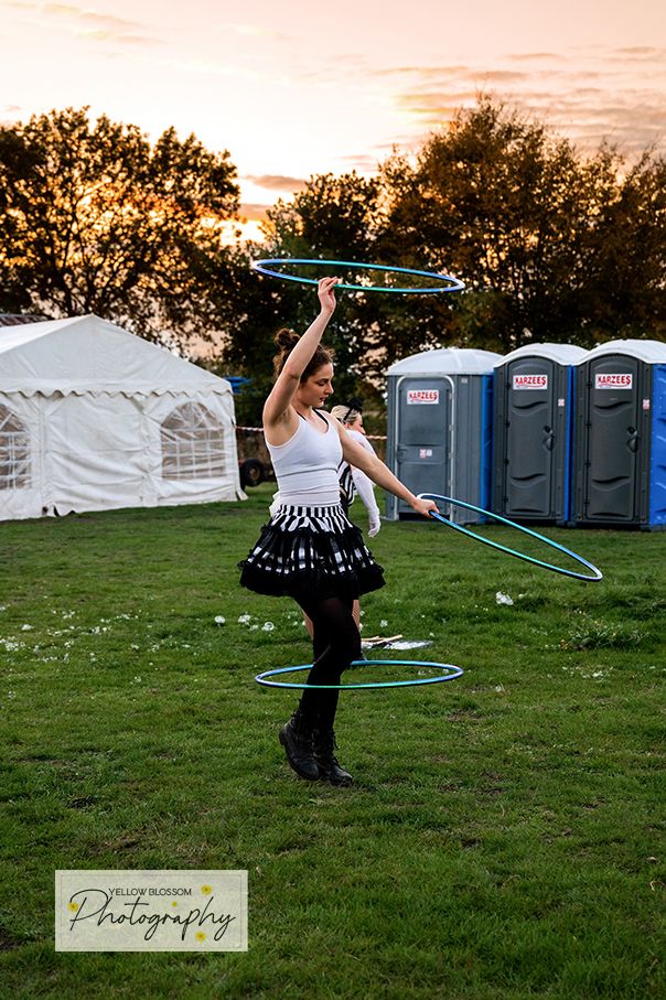 A performer in a black and white outfit spins a hula hoop on a grassy field with portable toilets in background.