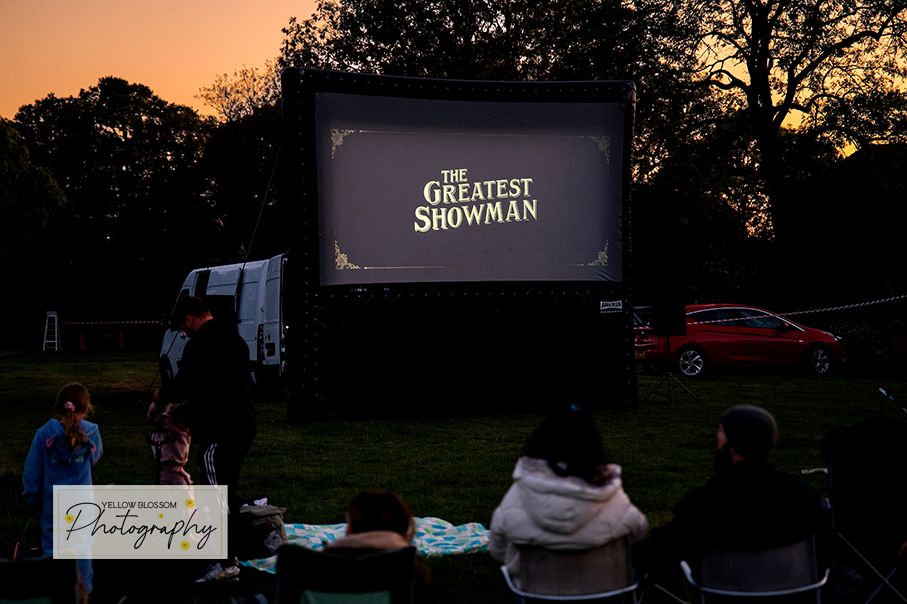 Silhouettes of trees against a sunset sky with outdoor movie screening setup visible in the background.