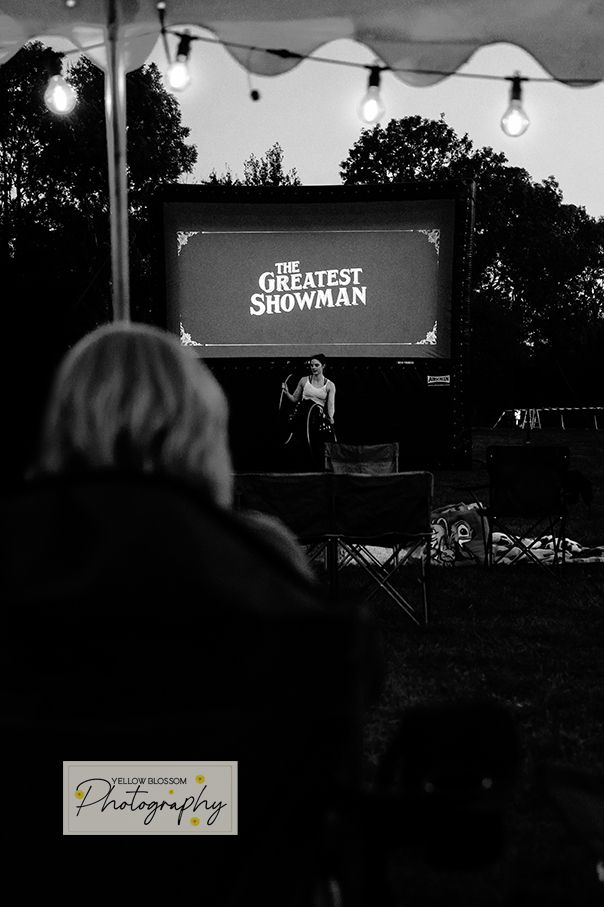 Black and white outdoor cinema setup with string lights and a movie screen showing 'The Greatest Showman' title.