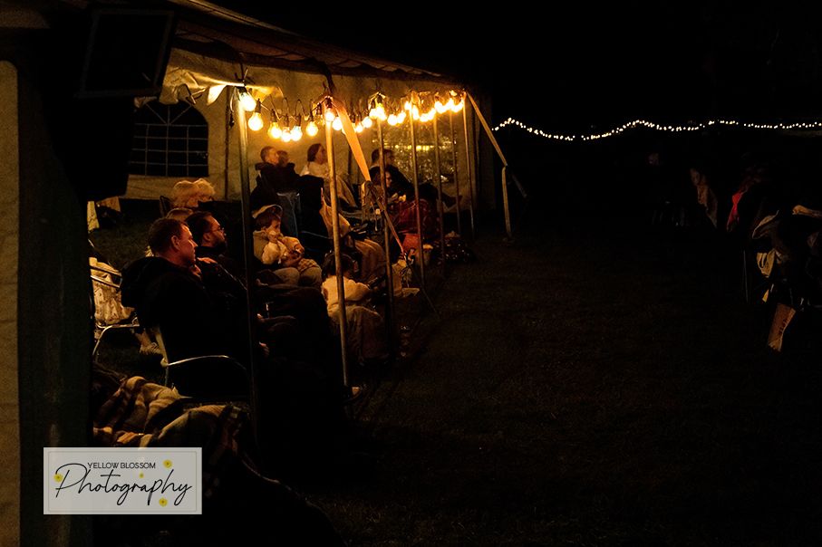 Guests gather under warm string lights on a dark porch during an evening outdoor celebration.