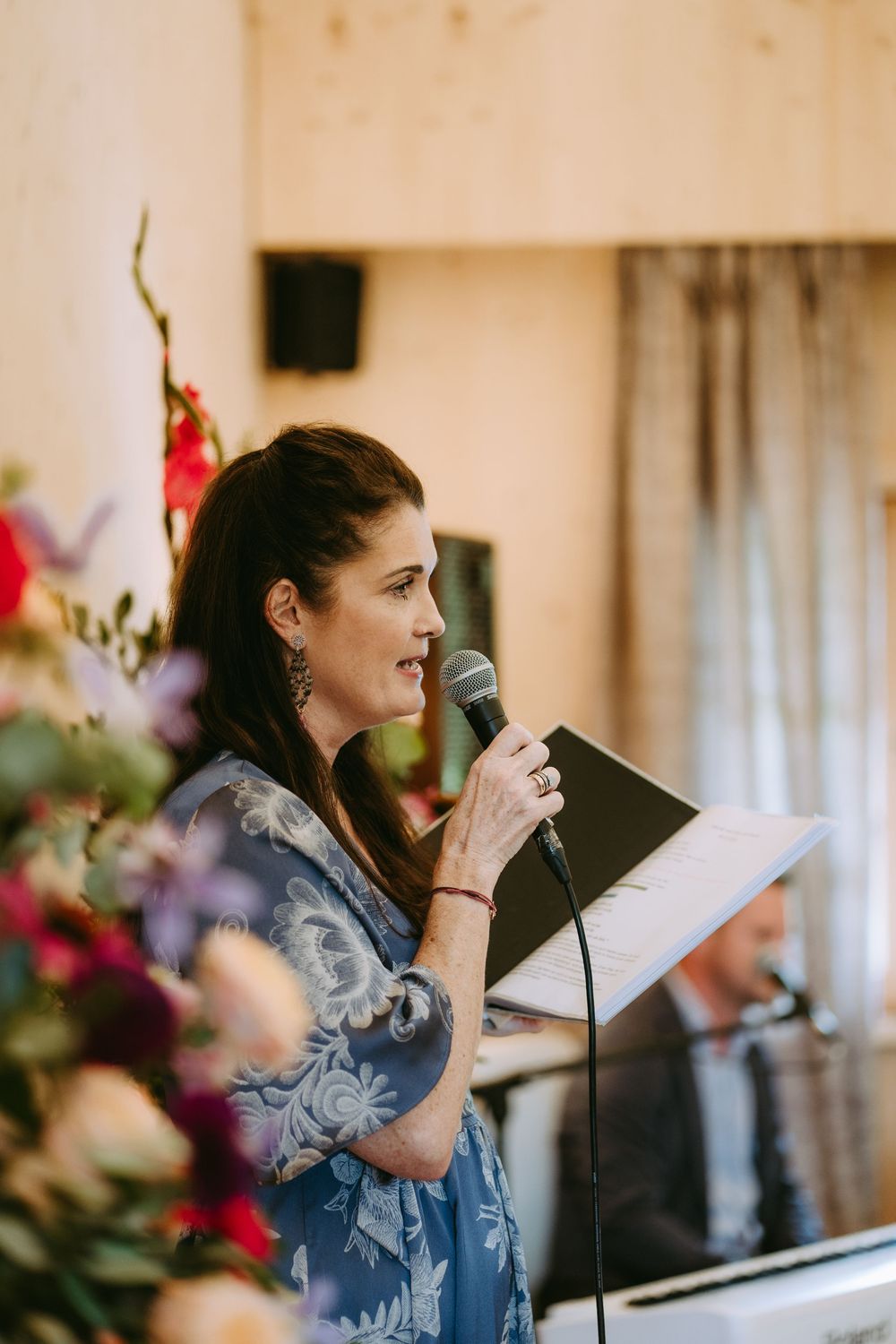 Speaker at wedding ceremony holding microphone next to floral arrangements.