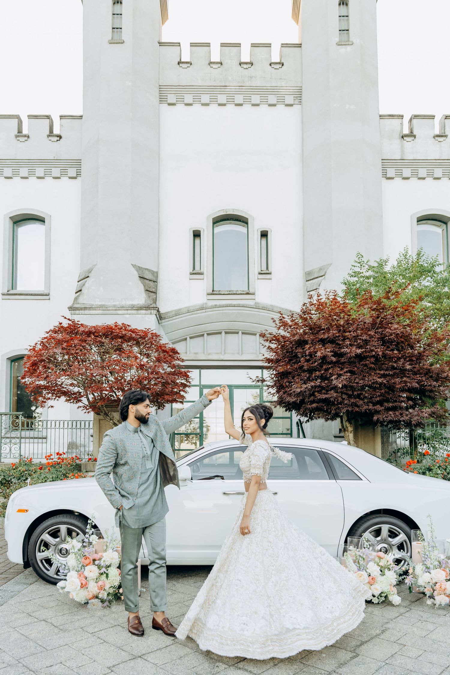 A wedding couple poses with their decorated white car in front of a castle-style building.