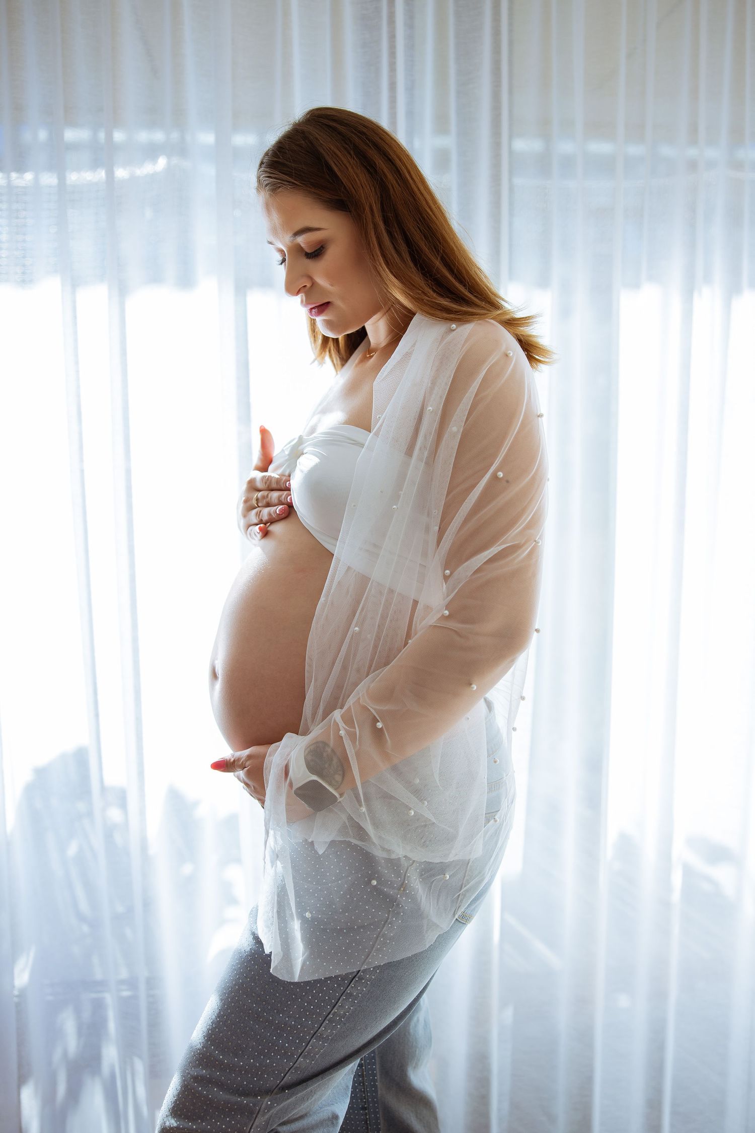Pregnant woman in white sheer top cradling baby bump by bright window during an at-home maternity shoot in Amsterdam.