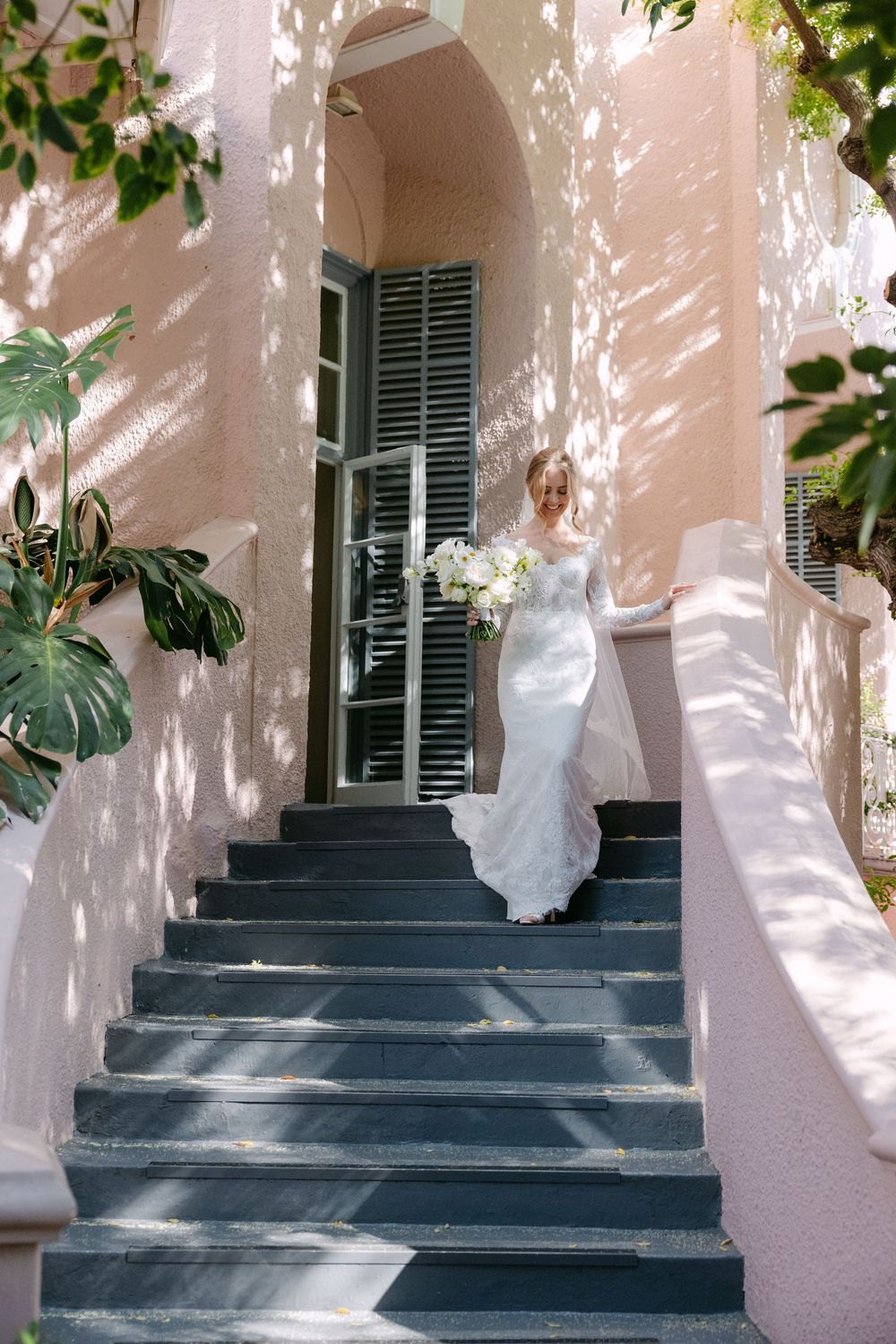 Sunlight casts dappled shadows on pink stucco walls and blue shutters along a Mediterranean-style staircase entrance.