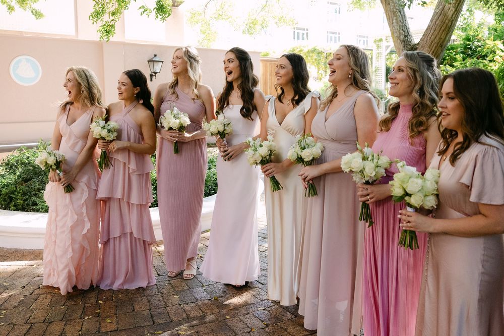 Bridesmaids in flowing pink and blush dresses stand together holding white floral bouquets during outdoor wedding.