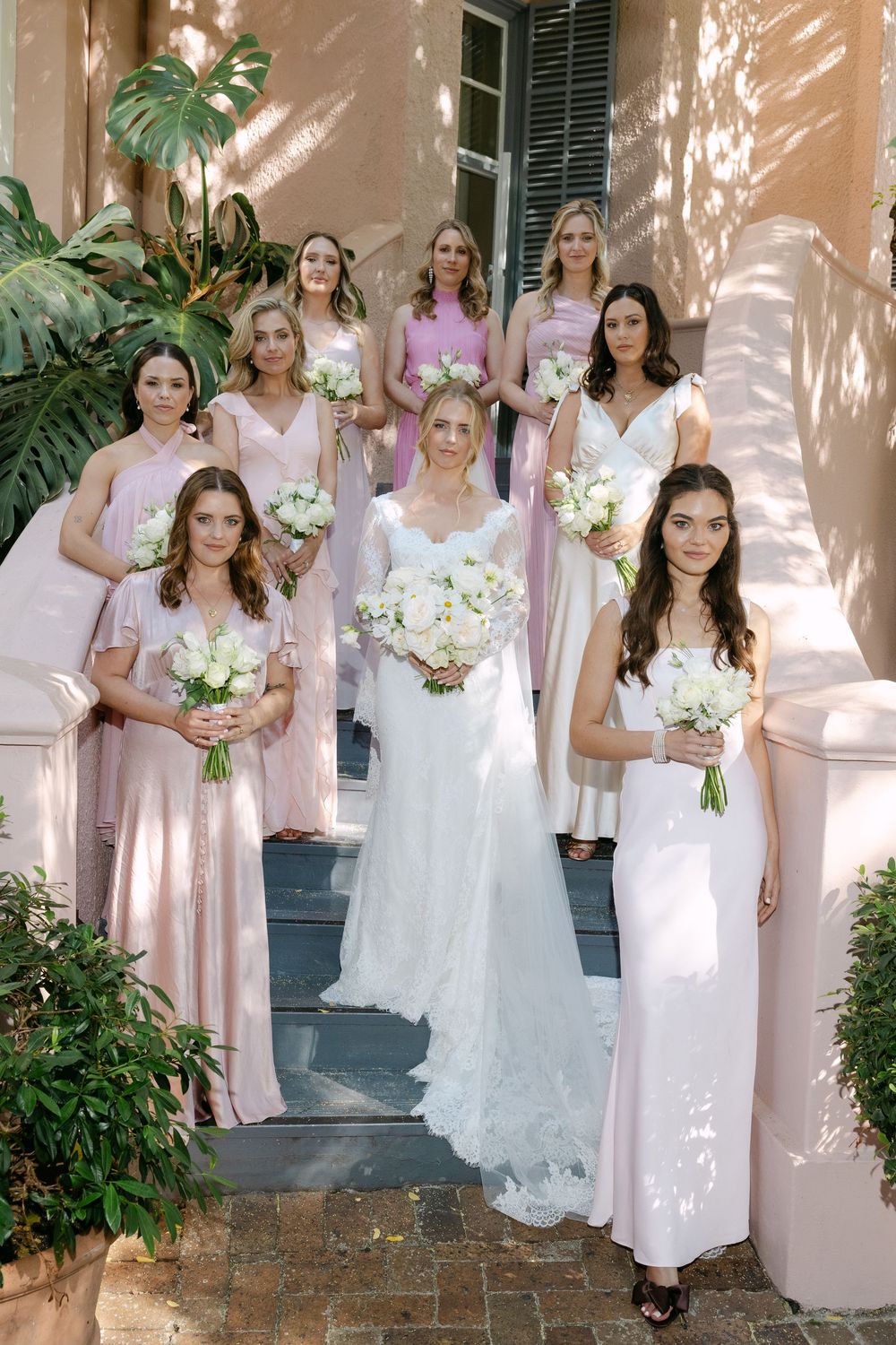 Wedding party in blush pink bridesmaid dresses and white bouquets pose on pink stucco steps with tropical greenery.