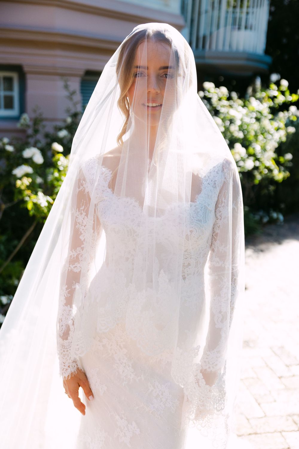 Elegant bride in a long-sleeved lace wedding dress with flowing veil stands in sunlit garden setting.