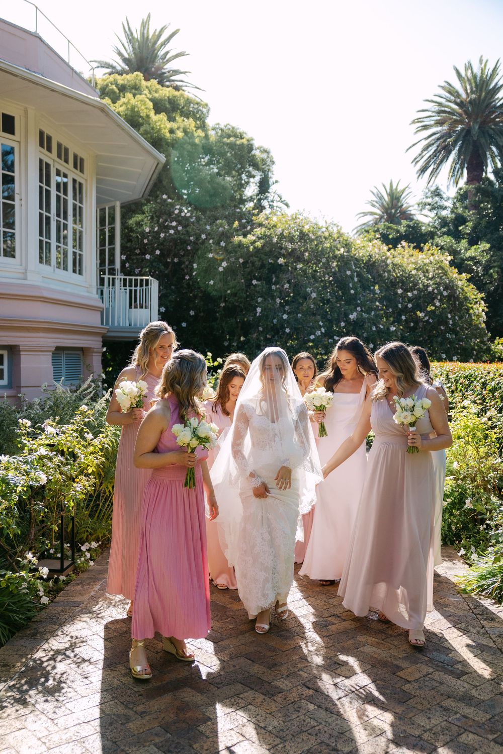 A bridal party in pink dresses walks along a sunlit brick path outside a white building with palm trees.
