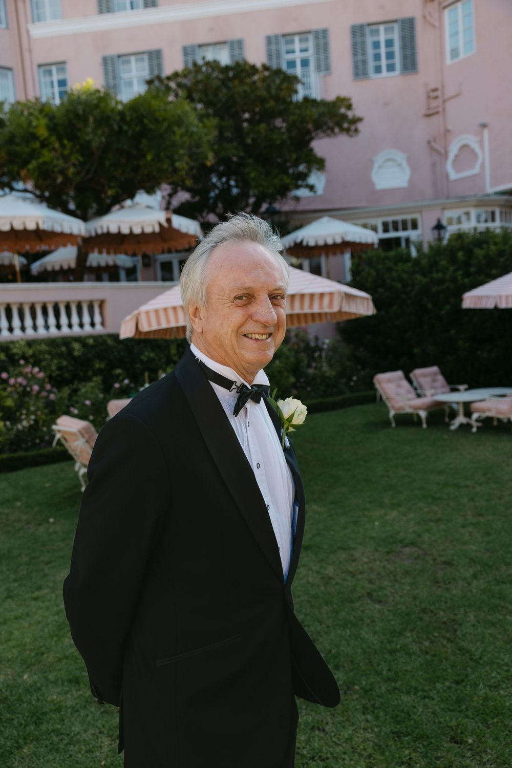 Formal wedding guest in classic black tuxedo smiles on lawn of pink hotel with umbrellas.