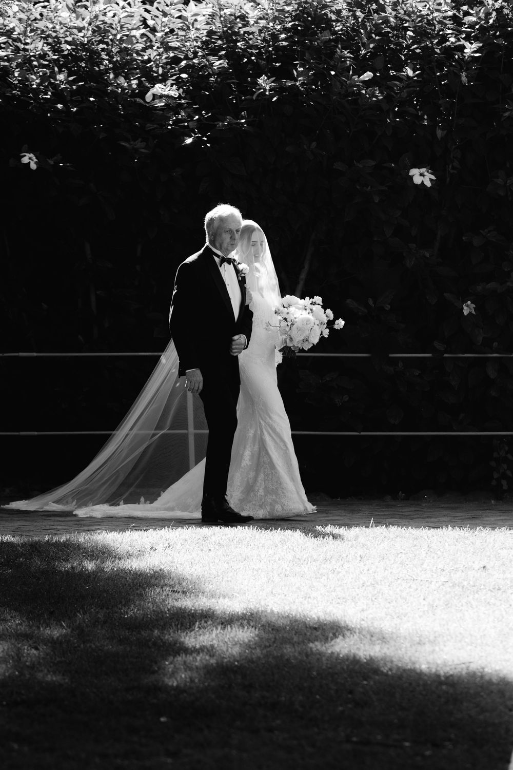 Black and white silhouette of bride and groom standing together with flowing veil in garden setting.