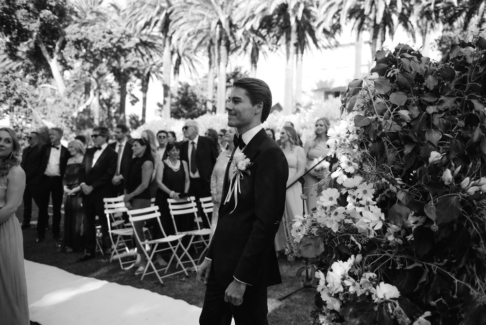 Wedding guests seated in white chairs at an outdoor garden ceremony surrounded by lush greenery and floral decor.