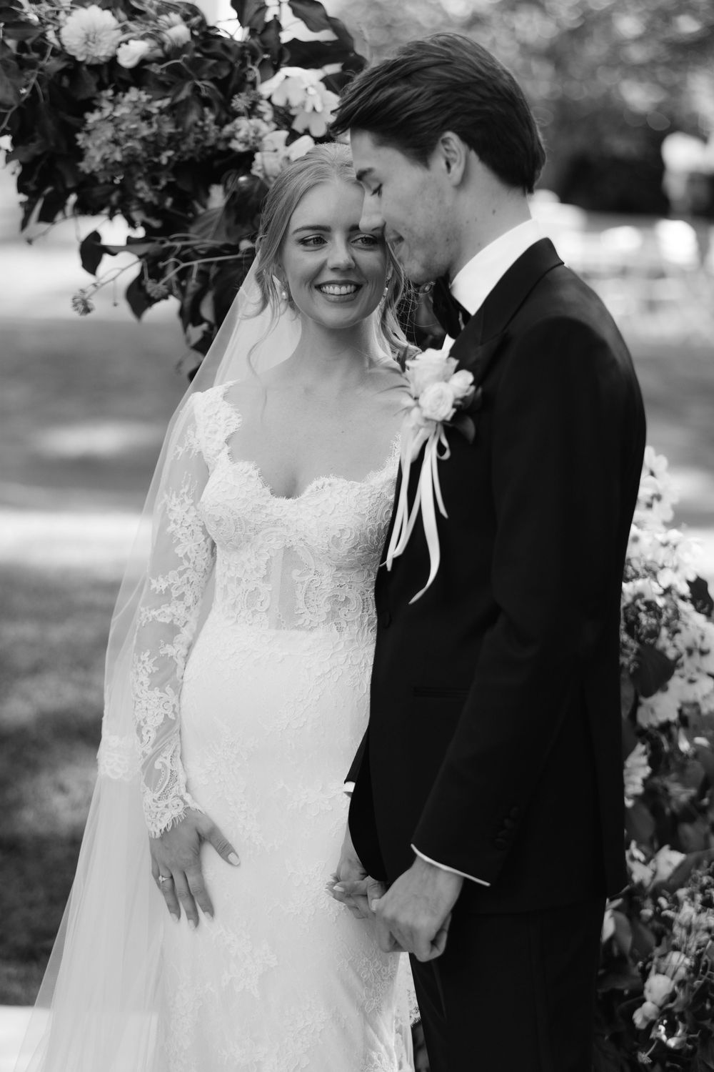 Black and white portrait of newlyweds sharing intimate moment under floral arch at outdoor wedding.