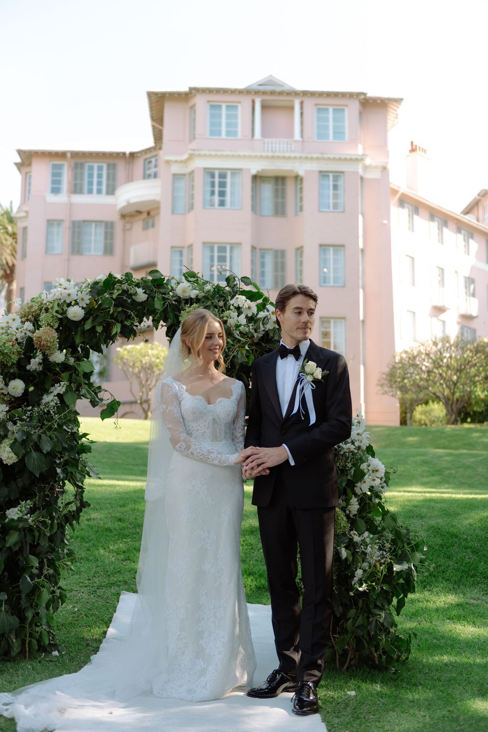 A couple celebrates their wedding day in front of a floral arch on a green lawn with a pink building in the background.