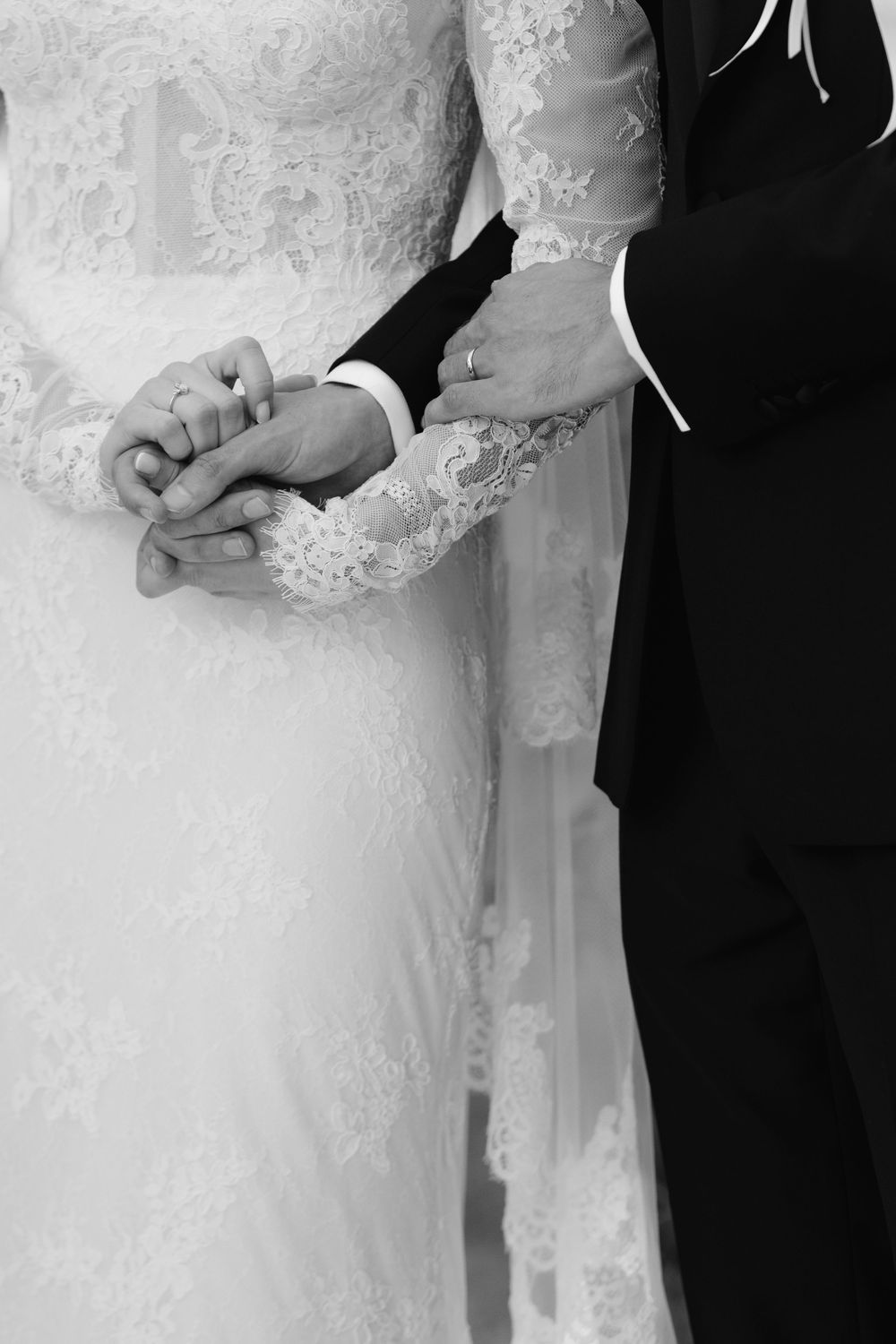 Black and white close-up of bride and groom's hands during wedding ceremony with lace dress detail.