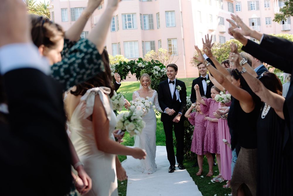 Wedding guests throw flower petals as newlyweds walk through an outdoor celebration path.