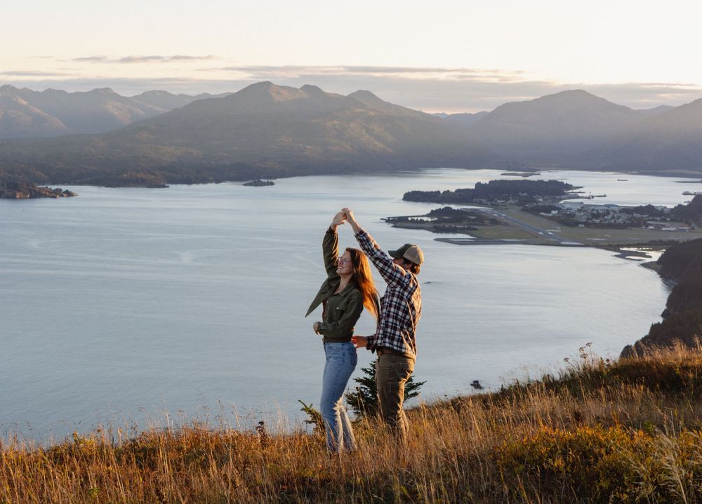 Panoramic view of coastal mountains and ocean bays during golden hour with hikers silhouetted against the sunset.